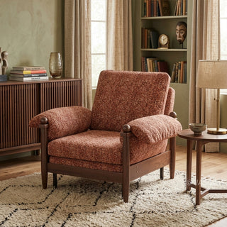 Lifestyle staging shot of a brown textured fabric accent armchair with dark wood framing in a cozy, book-filled living room.