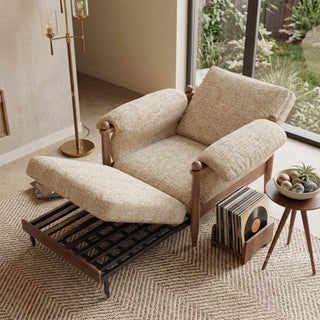 Lifestyle staging shot of a convertible accent armchair with its integrated pull-out footrest extended, featuring textured beige fabric and dark wood framing.
