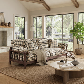 Three-quarter view of a modern plaid patterned sofa with a solid wood frame base, matching checkered accent throw pillows, and a cozy throw blanket in a sunlit living room with a round stone coffee table.