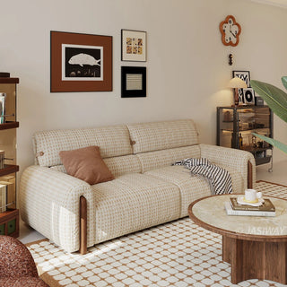 Modern cream and beige houndstooth tweed fabric sofa styled with a terracotta throw pillow and a patterned blanket, set in a cozy living room with art prints, a round wooden coffee table, and a flower-shaped wall clock.
