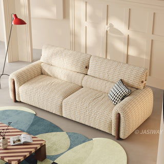 High-angle shot of a modern cream and beige houndstooth tweed fabric sofa with walnut wood trim accents, featuring a black and white plaid accent pillow on a geometric rug in a bright living room.