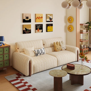 High-angle shot of a modern cream-colored houndstooth tweed fabric sofa with wood accents, styled with bunny cushions and patterned pillows, set against a gallery wall with 6 framed art prints in a cozy living room.