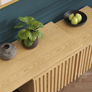 A high-angle close-up shot shows the surface of a modern light wood media console. The cabinet features distinctive vertical slatted paneling. On the surface, there is a potted green plant in a dark pot, a grey textured vase, and a black bowl filled with three pears.