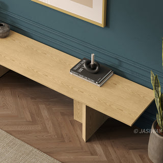 A high-angle close-up shot shows a section of a long light wood media console positioned against a dark teal wall with molding. On top of the console rests a stack of books with a black sculptural candle holder. A potted plant is visible on the right, and a herringbone wood floor is shown below.