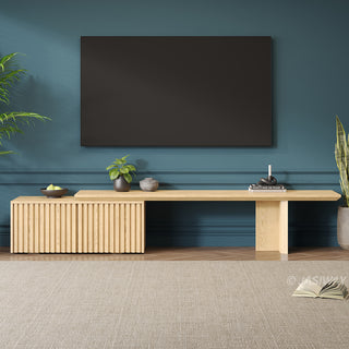 A large, dark television is mounted on a deep teal wall above a long wooden media console. The console features a slatted cabinet on one side and is decorated with small plants, a bowl of fruit, and books. Potted plants flank the setup, while a light-colored area rug sits on the wooden floor in the foreground.