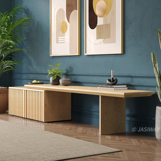 A modern light wood media console with a slatted cabinet is positioned against a deep teal wall. Above the unit, two framed abstract art prints feature geometric shapes in neutral and golden tones. Potted plants flank the console, while books and decorative items sit on its surface. A light-colored area rug rests on the herringbone wooden floor in the foreground.