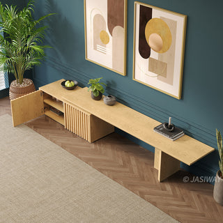 A long wooden media console is set against a dark teal wall, featuring an open cabinet door to reveal interior shelving. Above the unit, two framed abstract art prints hang on the wall, complementing the modern decor. Potted plants flank the setup, while a light-colored area rug rests on the herringbone wooden floor in the foreground.