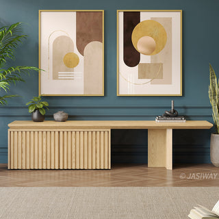 A modern light wood media console with a slatted cabinet is positioned against a dark teal wall. Above the unit, two framed abstract art prints feature geometric shapes in neutral and golden tones. Potted plants flank the console, while books and decorative items sit on its surface. A light-colored area rug rests on the herringbone wooden floor in the foreground.