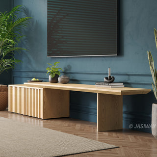 A long, light wood media console with a slatted cabinet is positioned against a deep teal wall. A large television hangs above the unit, reflecting a window with blinds. The console is styled with a small potted plant, decorative items, and a stack of books. Potted greenery flanks the console, and a textured rug rests on the herringbone wooden floor.