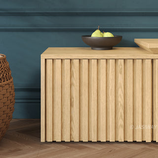A close-up shot highlights the vertical slatted paneling of a light wood media console. On top of the unit sits a dark bowl filled with green pears. The console is set against a dark teal paneled wall, with a woven basket and a herringbone wood floor visible nearby.