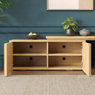 A light oak media console is centered against a dark teal wall with decorative molding. The console's cabinet doors are open, revealing four empty storage compartments, each with a circular cable management hole. The top surface displays a black bowl of pears and two vases with greenery, and a framed abstract art print is visible on the wall above. A light beige area rug is on the herringbone wood floor in the foreground.