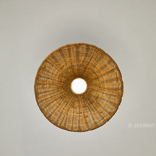 A centered, straight-up photograph of a circular woven rattan lampshade against a plain, light grey ceiling. The shade is composed of finely woven natural-colored rattan arranged in radiating spokes, with a single, glowing incandescent light bulb perfectly centered and visible within the central opening. The texture of the weave is clear, and the overall composition is minimalist and symmetrical.