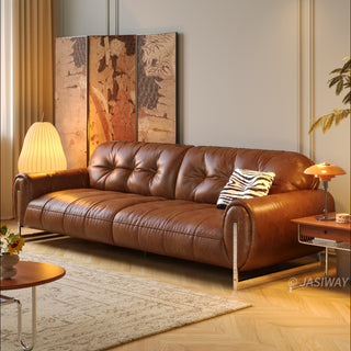 A rich tan leather sofa as the centerpiece of a warmly lit, stylish living room. The sofa boasts button-tufted back cushions and is accented with a zebra-print throw pillow, placed near a floor lamp, an ornate folding screen, and a side table. The space is completed with a patterned rug, herringbone wood flooring, and a small potted plant.