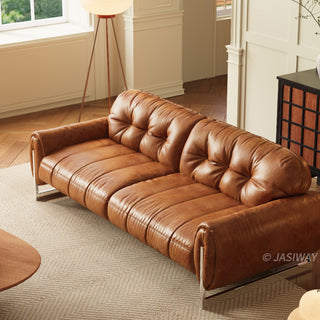 A high-angle shot of a luxurious, tan leather sofa with button-tufted back cushions and a smooth, paneled seat design. The sofa sits on a textured, light-colored rug in a bright room with herringbone wood flooring, near a floor lamp and a dark wooden cabinet.