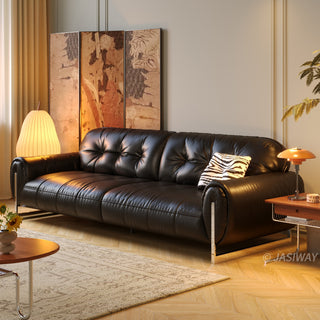 A sleek black leather sofa with button-tufted back cushions, serving as the focal point of a warm and stylish living room. It is accented with a zebra-print throw pillow and placed near an ornate folding screen, a contemporary lamp, and a wooden side table. The space is further enhanced by a textured area rug over herringbone flooring and ambient lighting.