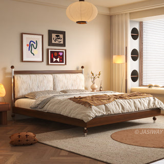 A serene, sunlit bedroom featuring a mid-century modern solid wood platform bed with a textured, neutral-toned upholstered headboard. The room is styled with patterned bedding, a decorative throw blanket, abstract wall art, and warm ambient lighting, including a large paper pendant lamp, with a cozy window nook featuring circular architectural details in the background.