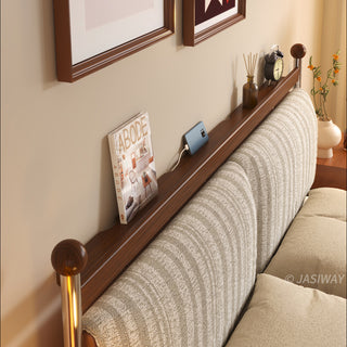A close-up, high-angle view of the functional headboard of a mid-century modern solid wood bed, featuring a built-in wooden ledge for storage that holds a magazine, a charging smartphone, and a small clock, positioned above a textured, striped upholstered headboard.