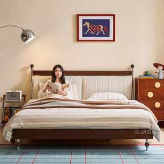 A model sits comfortably in a mid-century modern solid wood platform bed, reading a book. The bed features a textured, light-colored upholstered headboard and is styled with striped bedding. The bedroom setting includes a sleek floor lamp, a wooden dresser with circular accents, and a blue grid-patterned rug, completing the cozy interior design.