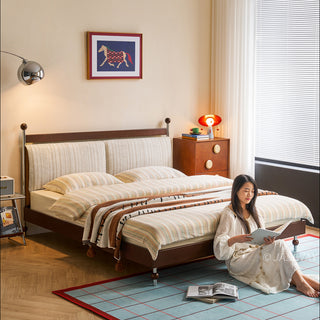 A model sits on a blue grid-patterned rug, reading a book in a sunlit bedroom featuring a mid-century modern solid wood platform bed with a textured upholstered headboard. The scene is styled with striped bedding, a wooden dresser with circular decorative accents, and a modern floor lamp, capturing a cozy, lived-in aesthetic.