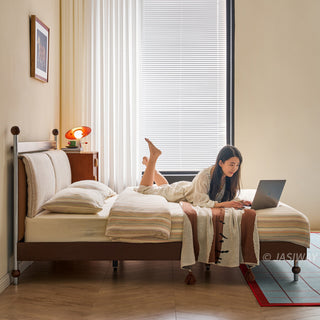 A model lying comfortably on a mid-century modern solid wood platform bed, working on a laptop. The bed features a textured upholstered headboard and is dressed in striped bedding with a decorative throw. The bedroom is bright and airy, with a large window covered by blinds, wooden flooring, and a blue grid-patterned rug.