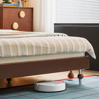 A white robotic vacuum cleaner operates efficiently under the clear space of a mid-century solid wood platform bed frame, which is equipped with distinct spherical feet. The scene includes the foot of the bed dressed with striped bedding and a fringed tassel, alongside a dark wooden dresser with circular knobs in the background, all resting on a blue grid-patterned rug.