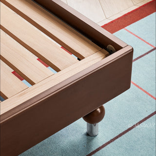 A detailed, close-up shot of the corner construction of a mid-century modern solid wood bed frame, showcasing the sturdy wooden support slats and the distinctive decorative spherical foot with a polished metallic accent, resting on a textured blue and red grid-patterned rug.