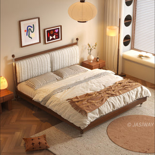 A high-angle interior view of a cozy bedroom featuring a mid-century modern solid wood platform bed with a textured upholstered headboard, styled with patterned bedding, a throw blanket, bedside tables, abstract wall art, and a sunlit window nook with circular storage accents, all set on herringbone flooring.