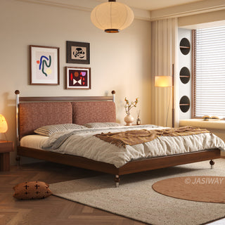 A warm, sunlit bedroom featuring a mid-century modern solid wood platform bed with a textured terracotta upholstered headboard, set against a wall with framed abstract art, a large paper pendant light, a cozy window nook, and herringbone wood flooring with layered rugs.