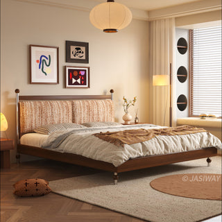 A wide-angle interior photograph of a warm, sunlit bedroom featuring a mid-century modern solid wood platform bed with a textured tweed-like upholstered headboard in a rust-orange hue, set on a herringbone wood floor with layered rugs. Abstract art is on the wall, a paper pendant light hangs from the ceiling, and a cozy window nook with circular storage built-ins is to the right.