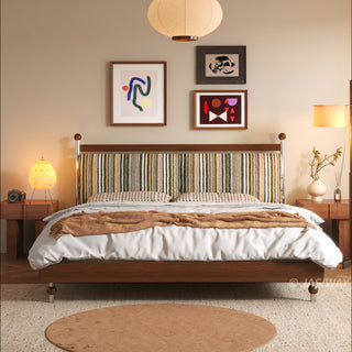 A frontal view of a mid-century modern solid wood platform bed featuring a vibrant, vertically striped upholstered headboard. The bed is styled with patterned bedding, a textured throw, and is framed by two wooden nightstands with soft accent lighting, a large paper pendant light above, and three abstract art pieces on the wall.