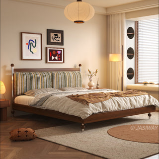 A serene, sunlit bedroom scene featuring a mid-century modern solid wood platform bed with a vibrant, vertically striped upholstered headboard. The room is detailed with herringbone wood floors, a cozy window nook with circular storage accents, framed abstract art, and warm, soft lighting from a large paper pendant lamp.