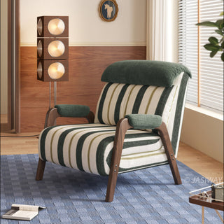 A mid-century modern armchair is upholstered in a textured fabric with broad vertical stripes in olive green, cream, and lighter green. The chair features a distinctive dark wood frame with angled legs and padded green armrests, along with a plush, solid olive-green headrest cushion. It sits on a blue patterned area rug, positioned in a room with wood paneling, a unique three-tiered spotlight floor lamp, a framed horse print, and a window with blinds.