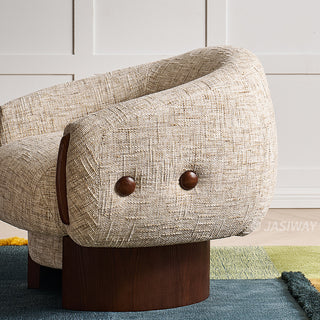 A side view highlights the textured beige fabric and curved silhouette of a modern armchair. The accent chair features dark wooden button accents on its side and a smooth wooden swivel base. It rests on a colorful, multi-toned rug against a clean white paneled wall.