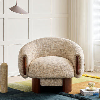A beige textured armchair with a curved, barrel back sits centered on a colorful multi-toned rug. The modern accent chair features dark wooden accents on its armrests and a sturdy wooden swivel base. To the right, a stack of books and a delicate teacup rest on the floor. A unique floor lamp stands to the left, set against a clean white paneled wall.