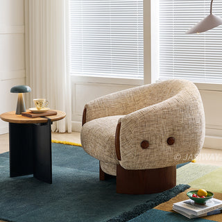 A beige textured armchair with a wooden base sits centered on a blue and yellow patterned rug. The modern accent chair features dark wooden accents on its armrests and buttons on the side. To the left, a small round wooden table holds a sleek lamp, a book, and a teacup. Large windows with white blinds fill the bright, cozy room with natural light.