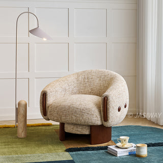 A beige textured armchair with a wooden base rests on a colorful patterned rug. The modern accent chair features dark wood accents on its curved armrests. To the left, a tall arched floor lamp stands against a white paneled wall. On the right, a stack of books, a teacup, and a small candle complete the cozy setup.
