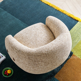 A high-angle, top-down photograph shows a round, textured beige armchair resting on a vibrant multi-colored rug. The rug features large, geometric sections of deep teal, yellow-green, and burnt orange. A small green bowl of lemons is visible on the teal section of the rug to the lower left. The chair has a distinctive U-shaped backrest and side buttons.