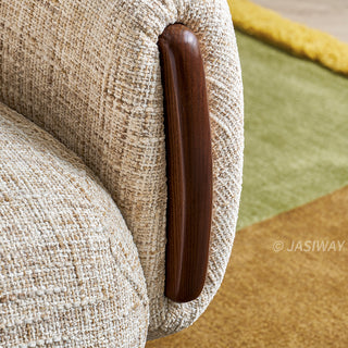 A close-up photograph focuses on the textured beige fabric of an armchair, showing intricate woven details. A vertical, smoothly polished dark wood accent is integrated seamlessly into the side of the armrest. The background features a colorful geometric rug with patches of green and gold. 