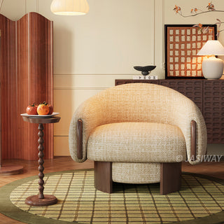 A beige textured armchair with dark wood accents on its curved armrests sits centered on a green patterned rug. To the left, a tall, slender wooden side table holds a small white plate with fruit. In the background, a dark wood cabinet is decorated with a lamp, a bowl, and abstract wall art, all set against a warm beige wall.