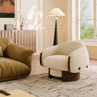 A textured beige armchair with dark wood accents on its armrests and side buttons sits on a light patterned rug. In the foreground, part of a larger olive-green sofa is visible to the left. A modern floor lamp and a wooden cabinet with a vase and radio decorate the bright room in the background.