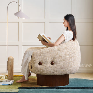 A profile view shows a model in a white dress sitting sideways in a textured beige armchair, focused on reading an open book. The armchair features dark wooden accents and a solid wood base, resting on a colorful multi-toned rug. To the left, a tall arched floor lamp stands beside a stack of books and a teacup, set against a clean white paneled wall.