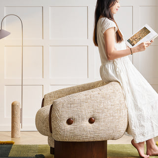 A model wearing a white dress sits on the back of a textured beige armchair while reading an open book. The chair features dark wooden button accents and a solid wood base, resting on a green rug. A modern floor lamp stands in the background next to a white paneled wall.