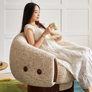 A model in a white dress sits comfortably in a textured beige armchair while holding a teacup and saucer. The modern accent chair features dark wood accents on the armrests and side buttons, adding a touch of elegance. Set against a clean white paneled wall, the cozy indoor scene exudes a calm and relaxing atmosphere.