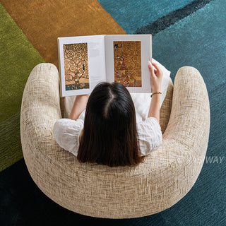 A top-down, high-angle view shows a model sitting from behind in a curved, textured beige armchair. The individual is wearing a white dress and is holding open an art book with colorful illustrations. The modern armchair rests on a vibrant multi-toned rug featuring patches of green, brown, and teal.