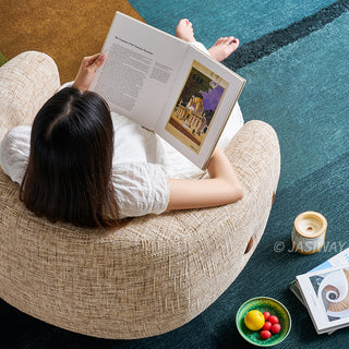 A top-down view shows a model sitting in a textured beige armchair while reading an open book. The individual is wearing a white dress and has their bare feet resting near the top of the chair. The chair is placed on a multi-colored carpet next to a small bowl of fruit and stacked books.