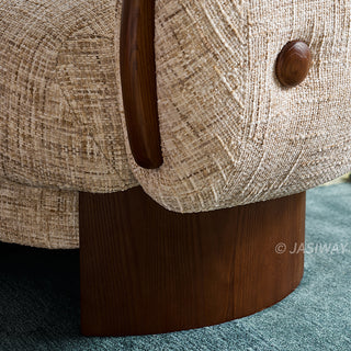 A close-up view highlights the textured beige fabric and dark wooden details of a modern armchair. A smooth dark wood armrest curve and a decorative wooden button accent the side. The chair is supported by a sturdy, curved dark wood base resting on a blue carpet.