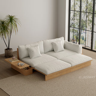 A wide, low-profile modular sofa bed with a natural wood frame is extended in a brightly lit room next to a large window. The thick cushions and scatter pillows are upholstered in a light beige fabric. An attached side table features a built-in drawer holding small items, with books and headphones resting on top. A potted palm tree stands beside the sofa on a textured beige area rug.