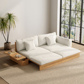 A wide, low-profile modular sofa bed with a natural wood frame is extended in a brightly lit room next to a large window. The thick cushions and scatter pillows are upholstered in a light beige fabric. An attached side table features a built-in drawer holding small items, with books and headphones resting on top. A potted palm tree stands beside the sofa on a textured beige area rug.