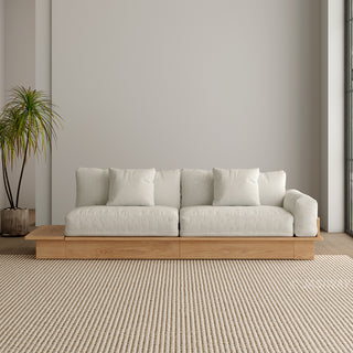 A modern, low-profile modular sofa with a natural wood base is positioned against a neutral grey wall. The thick cushions and matching scatter pillows are upholstered in a light cream fabric. An extended wooden platform extends from the left side of the sofa base. A potted palm plant stands to the left, and a textured beige area rug covers the floor in front.