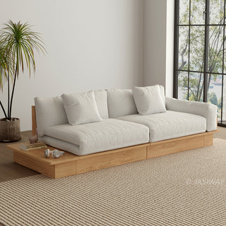 A wide, low-profile modular sofa with a natural wood frame sits in a bright room next to a large window. The thick cushions and scatter pillows are upholstered in a light beige fabric. An extended wooden platform acts as a side surface, holding small books and headphones. A potted palm tree stands beside the sofa on a textured beige area rug.